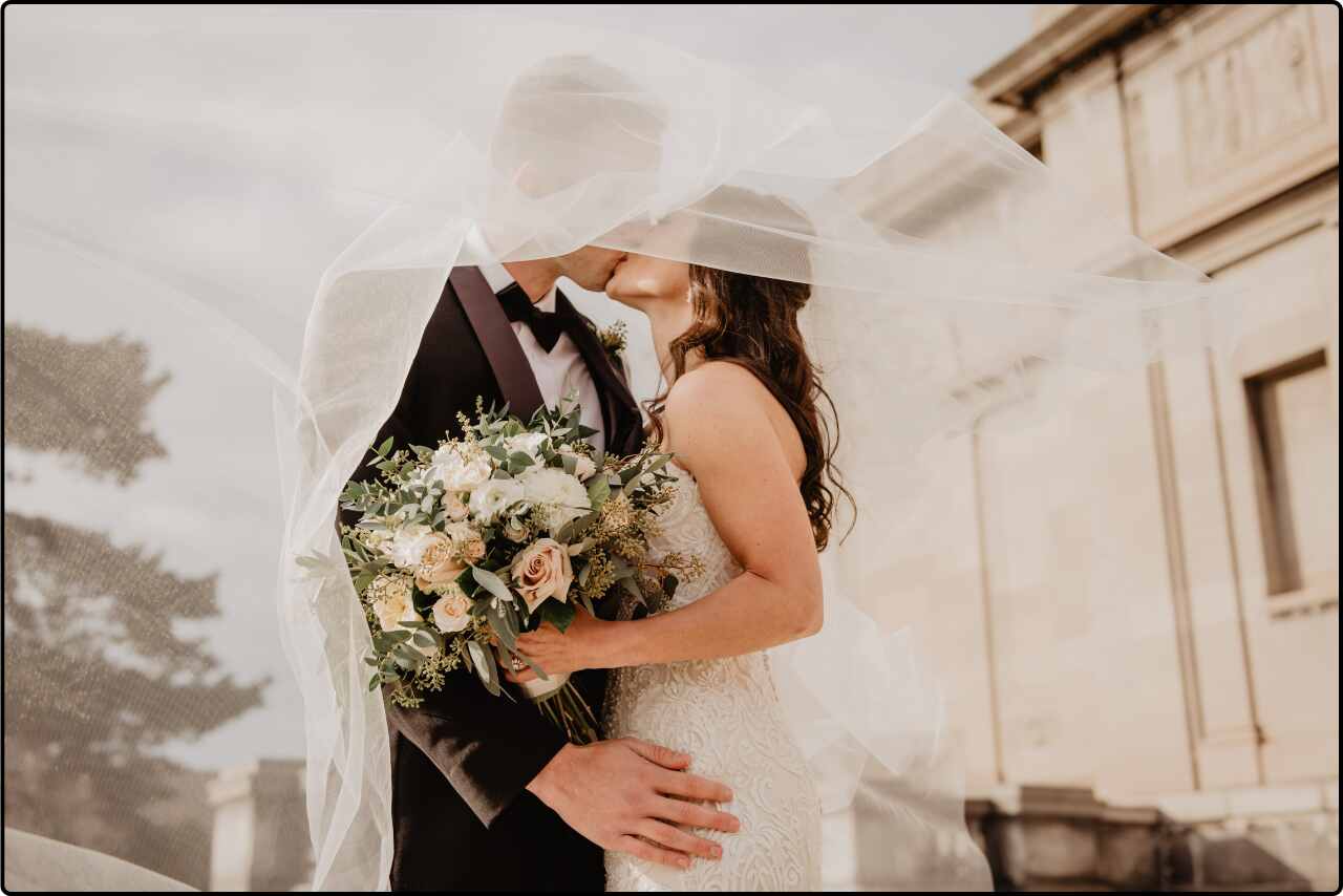 Bride and groom share a romantic kiss under a dress during their outdoor wedding ceremony